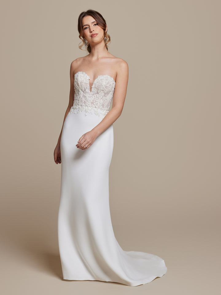 A woman wears the Esme wedding dress by averyaustin in Ivory/Nude Glitter, featuring a strapless sheath silhouette with a fitted, beaded floral lace bodice and smooth skirt, standing against a plain beige background.
