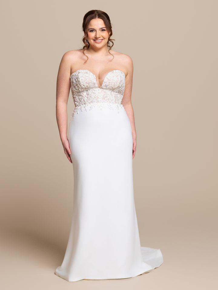 A woman smiles against a beige background wearing the Esme by averyaustin, a strapless sheath wedding dress in Ivory/Nude Glitter with a fitted, beaded floral lace bodice and a flowing skirt.