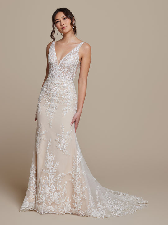A woman wears the Gabrielle by averyaustin, a sleeveless, floor-length ivory/nude lace wedding dress with a deep V-neckline and floral embroidery, posing against a beige background with one hand by her side and the other touching her gown.