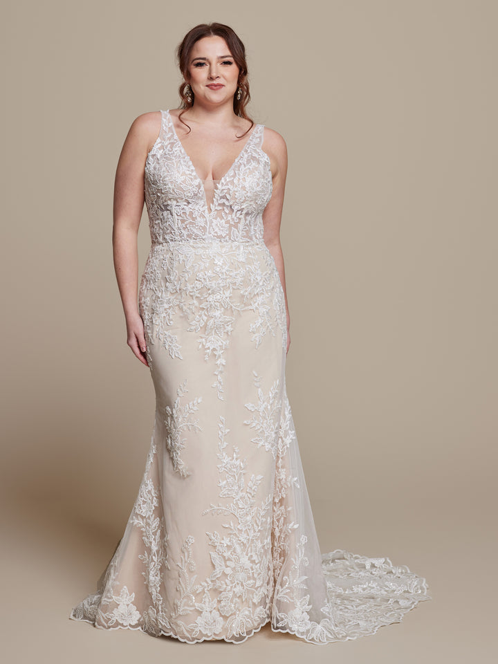 A woman smiles softly in the Gabrielle wedding dress by averyaustin, featuring a deep V-neck, intricate ivory floral lace over a nude underlay, and a sleeveless, floor-length silhouette that highlights her brown, wavy hair.