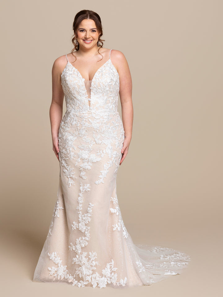 A woman smiles in the Vivienne by averyaustin, an ivory/nude glitter fit-and-flare wedding dress with beaded lace floral patterns, sleeveless design, and long train, posing against a plain beige background.
