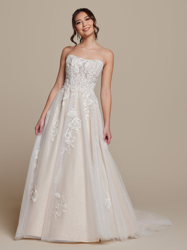 A woman smiles in the averyaustin Simone wedding dress, an ivory/nude glitter strapless corset A-line gown with beaded floral lace appliqué and subtle shimmer, elegantly displayed against a neutral beige background.