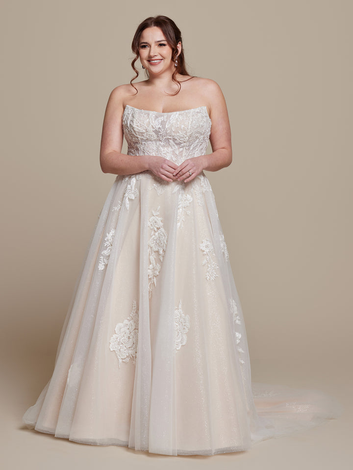 A woman smiles in the Simone wedding dress by averyaustin, featuring a strapless fitted bodice with beaded floral lace, a full tulle skirt with a subtle train, and an Ivory/Nude Glitter finish. Her brown hair is styled in loose waves against a beige background.