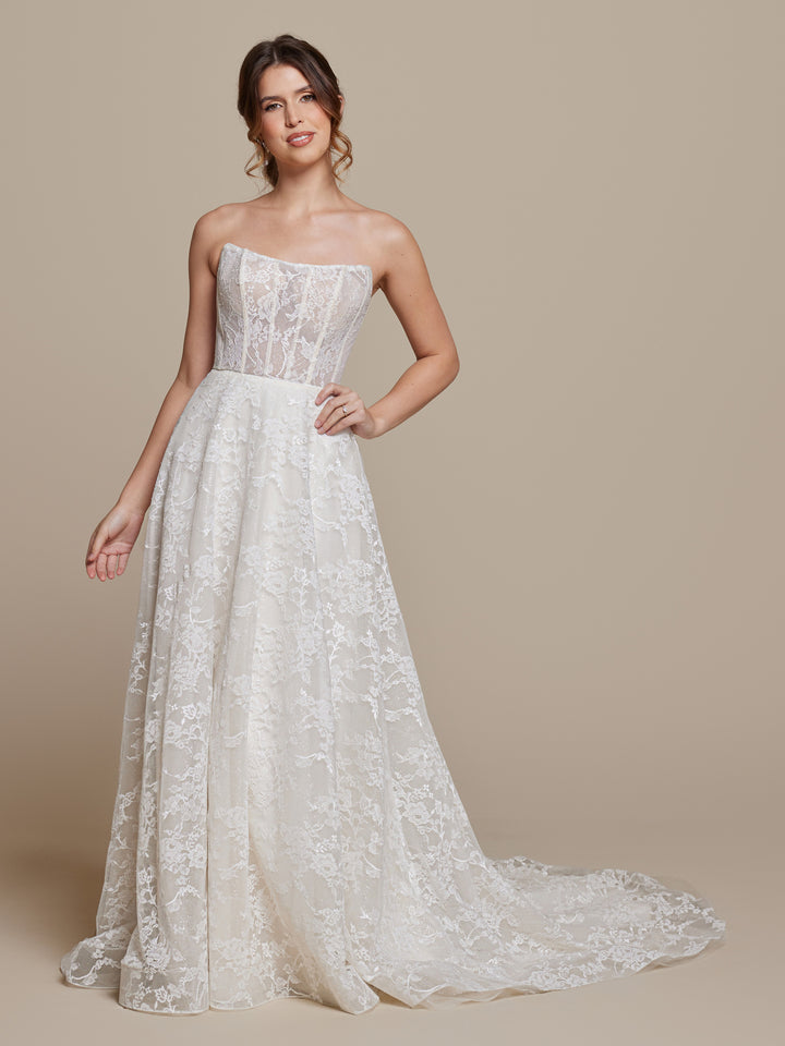 A woman in the Anais wedding dress by averyaustin, featuring a strapless fitted bodice and flowing ivory/nude glitter tulle and lace skirt, stands against a plain beige background, smiling softly.