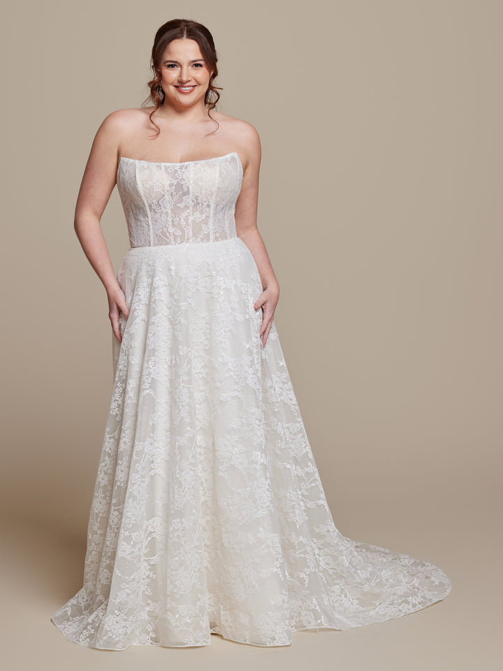 A woman wearing the Anais wedding dress by averyaustin in Ivory/Nude Glitter, featuring a strapless A-line silhouette with a lace semi-sheer bodice and long flowing skirt, stands smiling against a beige background.