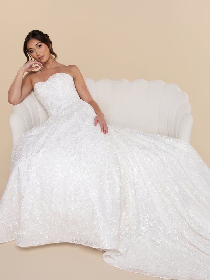 A woman wearing the ivory strapless A-line Celeste wedding dress with a floral pattern sitting on a scalloped-back sofa.