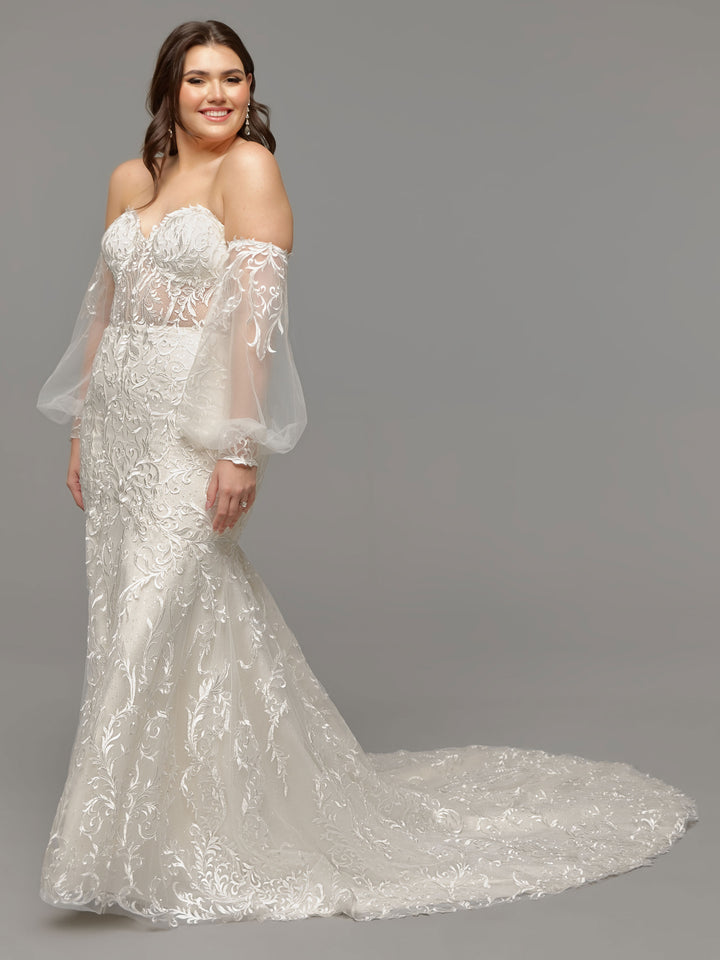 A woman in the Avery Austin TH-Kalea wedding dress—an ivory fit-and-flare gown with an off-the-shoulder lace bodice, sheer billowy sleeves, and a flowing train—stands against a gray background, smiling gently.