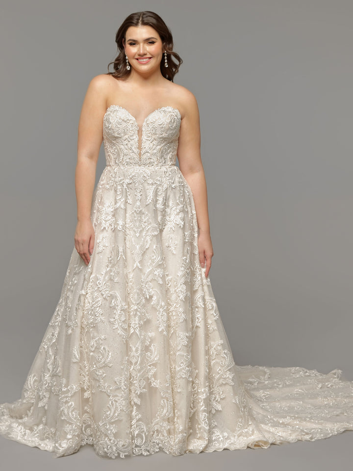 A woman smiles in the Alani by averyaustin, an ivory/sand strapless lace wedding dress with a sweetheart neckline, floor-length silhouette, intricate lace details, and a long train against a plain grey background.