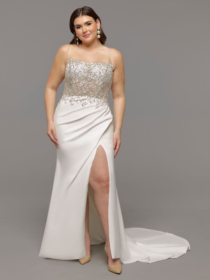 A woman poses confidently in the Avery Austin TH-Kiana ivory wedding dress, featuring a sheer embellished bodice, thin straps, fit-and-flare silhouette with a high slit and small train. She pairs it with drop earrings and beige heels against a gray background.