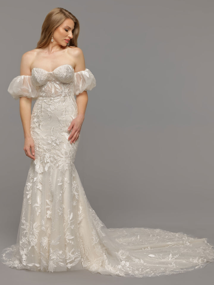 A woman wears the Avery Austin TH-Leilani, an ivory/ivory off-the-shoulder, fit-and-flare wedding dress with puffed sleeves and a long train, standing against a plain gray background.
