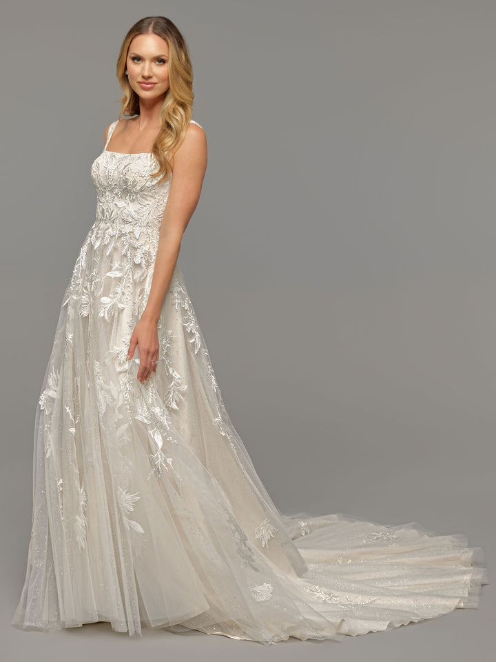 A woman models the Avery Austin TH-Luna wedding dress in Ivory/Ivory—a floor-length, open-back gown with sequined lace and a long train—against a gray background. She has long wavy blonde hair and smiles softly at the camera.