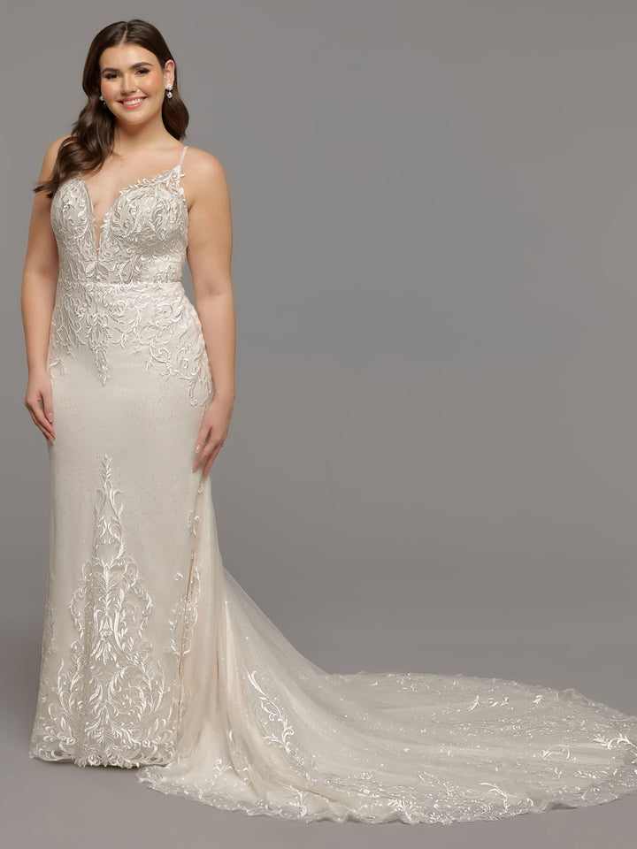 A woman poses against a gray background in the Avery Austin TH-Malia, an ivory/ivory fit-and-flare wedding dress with lace detailing, a plunging neckline, and an elegant train.