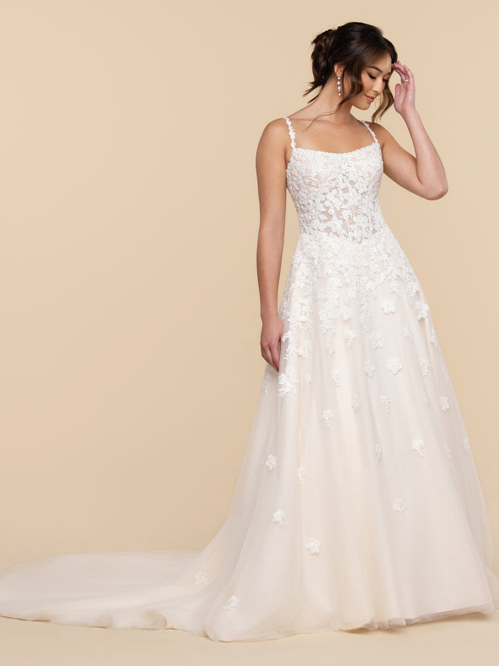 A woman wears the Miasie A-line wedding dress by averyaustin, featuring a square neckline, thin straps, and a skirt covered in lace and tulle floral appliqués.