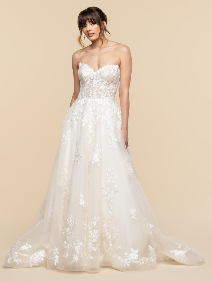 Liana by averyaustin is a strapless ivory and nude A-line wedding dress with a sweetheart neckline, sheer lace bodice, and full tulle skirt featuring floral appliqué and a long train.