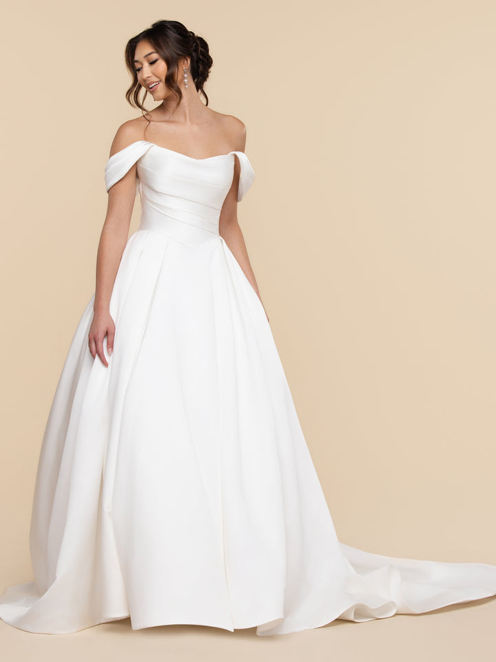 A woman wearing the Sage ivory off-the-shoulder satin ball gown with a full, floor-length skirt and a train.