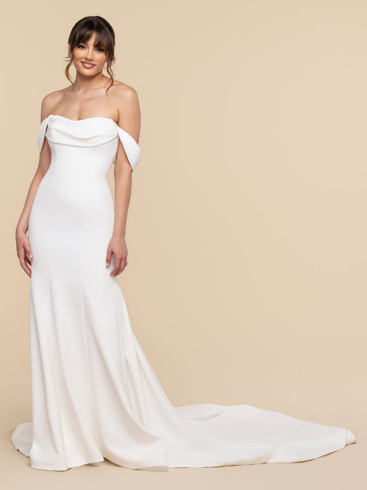 The Romy wedding gown by averyaustin features a romantic white off-the-shoulder draped neckline, a floor-length skirt, and a train.