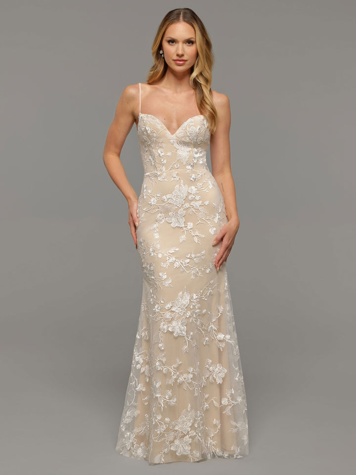 A woman poses in the Avery Austin TH-Maris gown, a fitted, floor-length ivory/nude wedding dress with white floral lace overlay, thin spaghetti straps, and a subtle v-neck, set against a plain gray background.