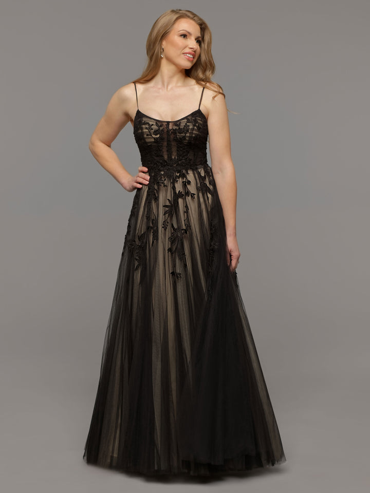 A woman in the Avery Austin TH-Kendall floor-length gown in Black/Nude with spaghetti straps and lace appliques stands against a plain gray background, smiling and gazing to the side.