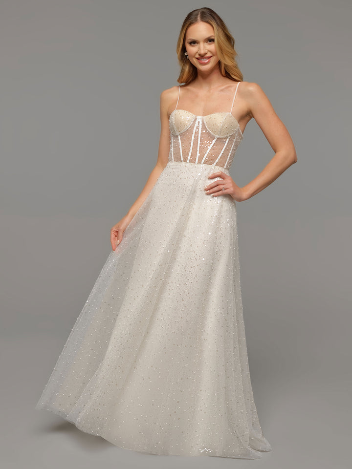 A woman in the Avery Austin TH-Margot, a sparkling floor-length beaded lace wedding dress with sheer panels, spaghetti straps, and a fitted bodice, smiles and poses against a plain gray background, holding the skirt.