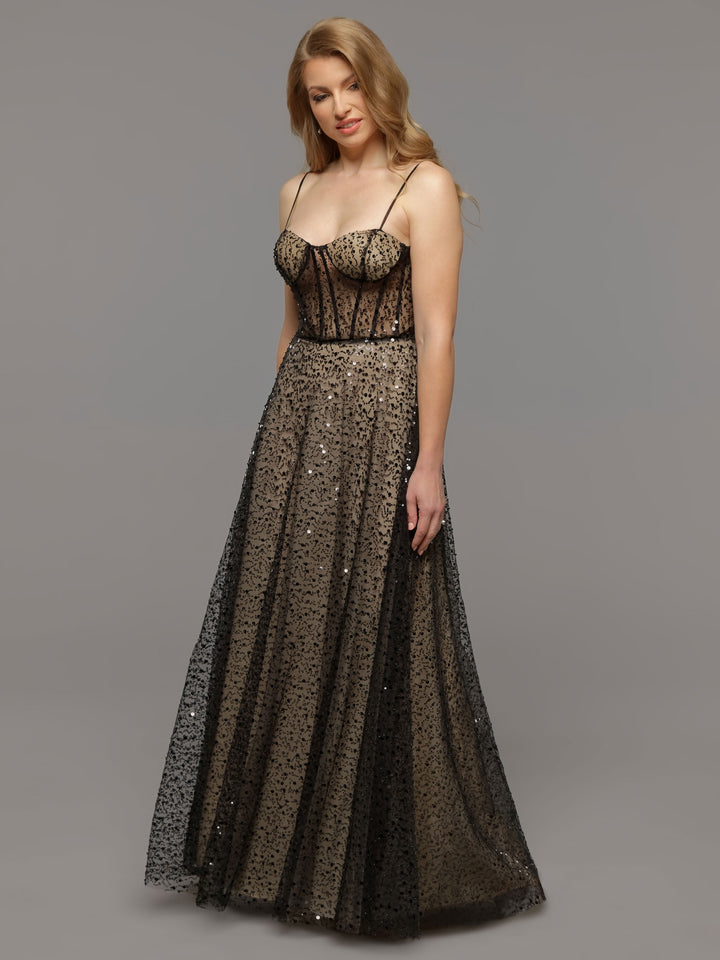 A woman models the Margot gown by averyaustin in Black/Nude, featuring a long, sleeveless silhouette with sequin details and sheer overlay, standing against a gray background with a poised, relaxed expression.