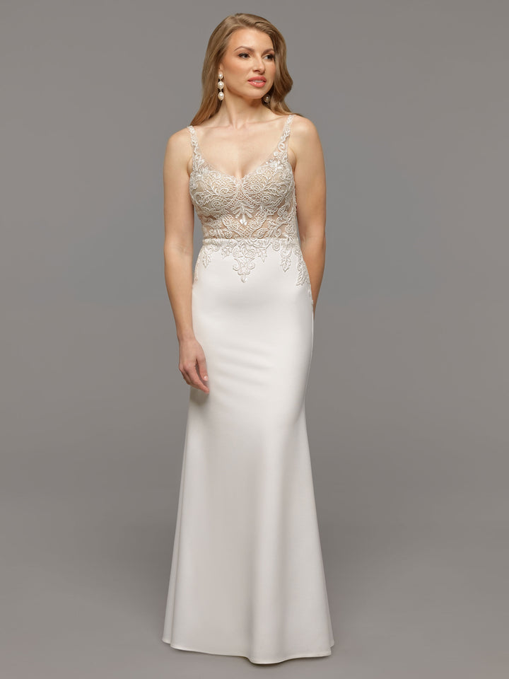 A woman wears the Avery Austin TH-Lilo gown in Ivory/Nude, featuring intricate lace bodice detailing and a fitted, floor-length skirt. Against a gray backdrop and accentuated with drop earrings, she exudes elegant, wedding-inspired style.