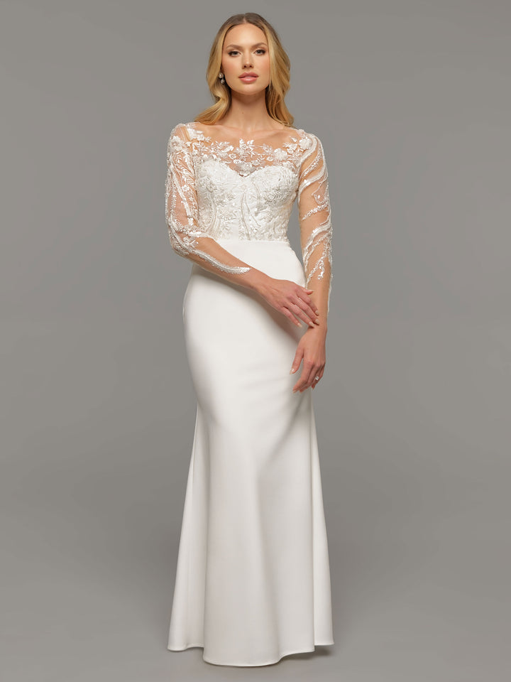 A woman stands against a plain gray background wearing the Avery Austin TH-Reese ivory wedding dress, featuring sheer embroidered long sleeves and a fitted bodice.