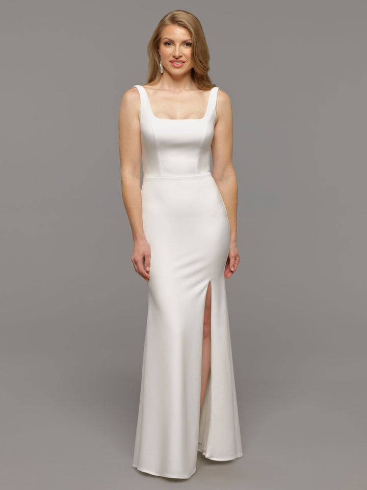 A woman smiles at the camera, wearing the Haven by averyaustin—a sleeveless, fitted white bridal gown with a square neckline and high front slit—standing against a plain gray background.