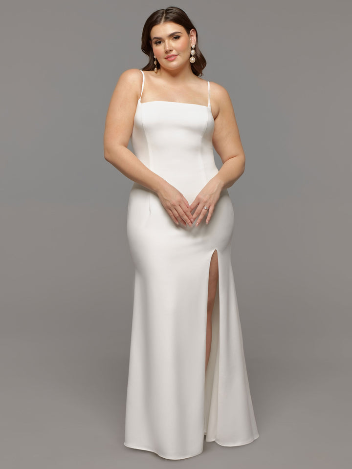 A woman in the Alora by averyaustin, a modern white fit-and-flare wedding dress with thin straps and a thigh-high slit, stands against a gray background. Her wavy brown hair and statement earrings complete her elegant look.