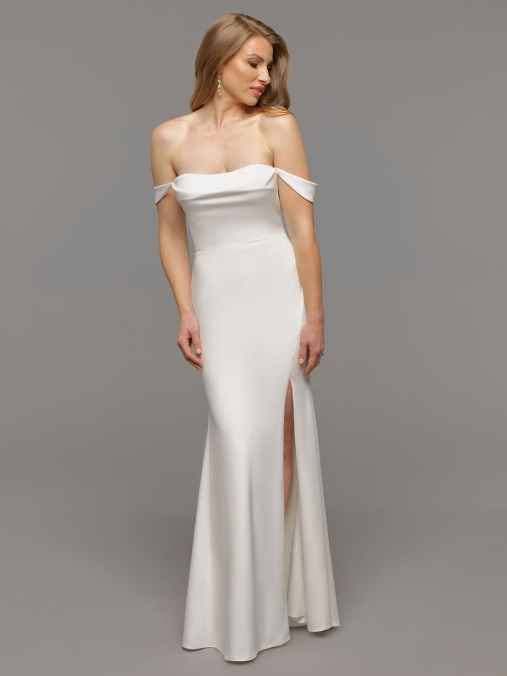 A woman models the Avery Austin TH-Evie wedding dress in ivory, featuring an elegant off-the-shoulder design and front slit, standing against a plain gray background and gazing slightly to her left.