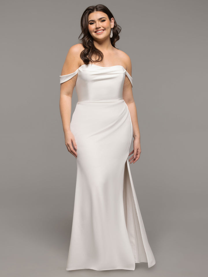 A woman stands and smiles gracefully against a plain grey background, wearing the Avery Austin TH-Evie wedding dress in ivory—an off-the-shoulder gown with a fitted bodice, slight slit, and floor-length skirt.
