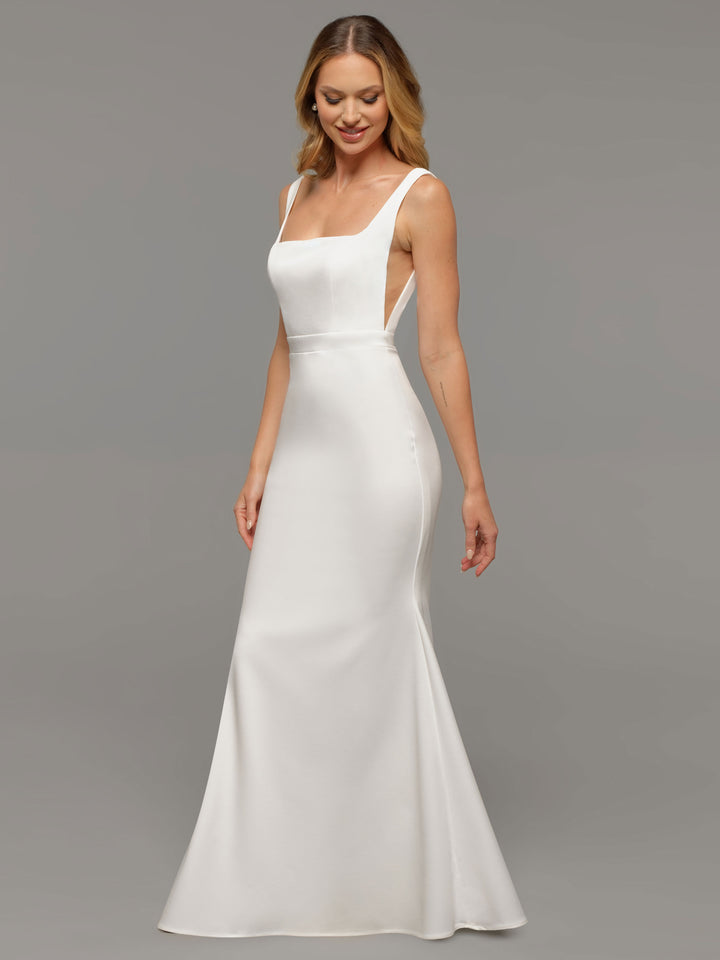 A woman wears the Tessa wedding dress by averyaustin—a sleeveless, floor-length gown with a square neckline and fitted silhouette—standing against a plain gray background, smiling slightly while looking down.