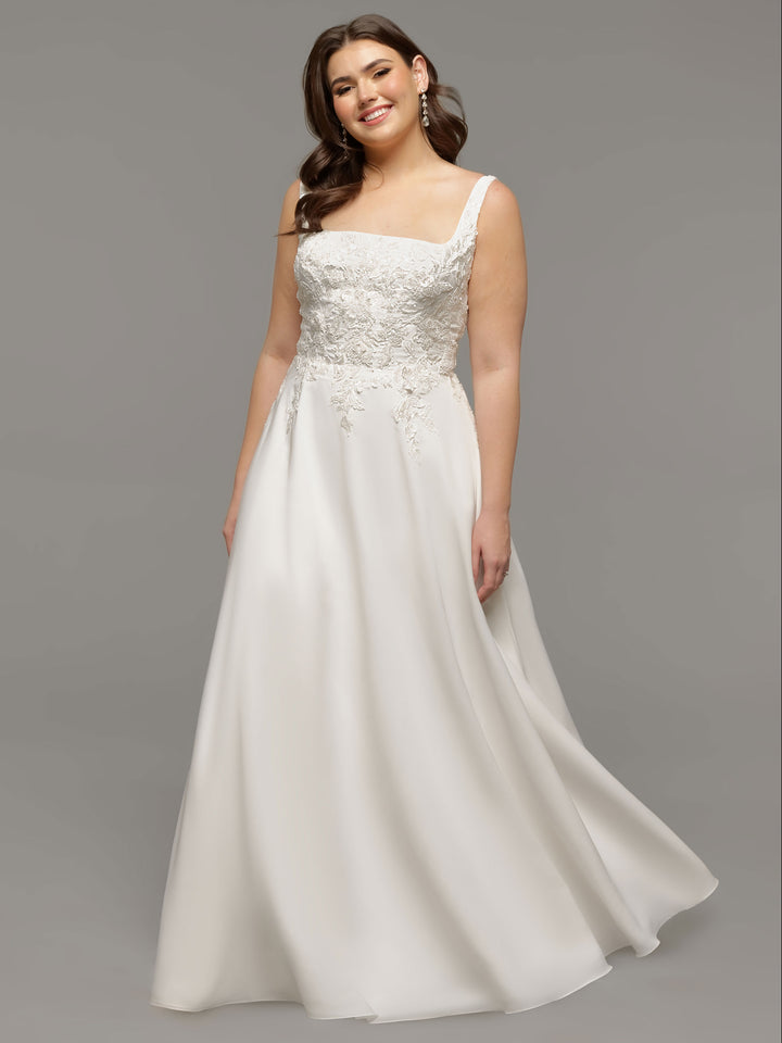 A woman smiles against a gray background wearing the Avery Austin TH-Scarlett wedding dress in ivory—a sleeveless, A-line gown with a square neckline and floral lace bodice, holding the skirt with one hand.