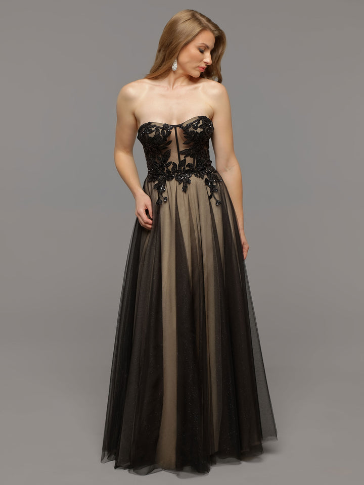 A woman in the averyaustin Phoenix gown in Black/Nude, featuring a strapless sheer design with floral lace appliques and a flowing skirt, looks down while standing against a plain gray background.