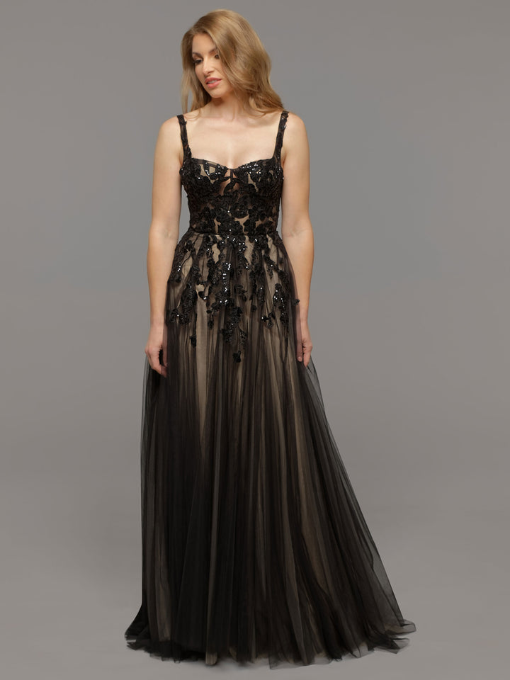 A woman stands against a gray background wearing the Coco gown by averyaustin in Black/Nude, featuring a floor-length, sleeveless design with a sheer tulle skirt and intricate black floral embellishments on the bodice and upper skirt.