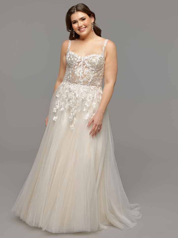 A woman smiles against a gray background wearing the Avery Austin TH-Coco wedding dress in Ivory/Nude, featuring a sleeveless, sheer lace bodice with floral appliqués and a flowing layered tulle A-line skirt.