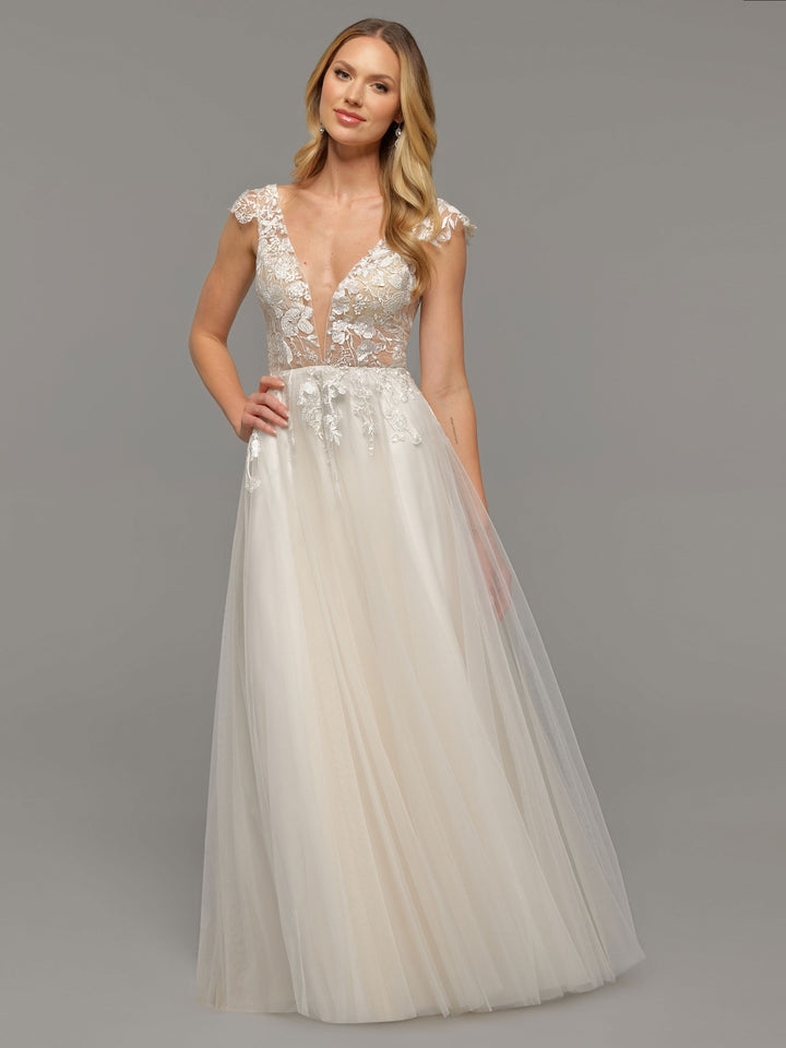 A woman wears the Avery Austin TH-Hana wedding dress in Ivory/Ivory, featuring a sleeveless A-line silhouette with a sheer deep V-neck lace bodice and flowing tulle skirt, standing against a plain gray background and smiling softly.