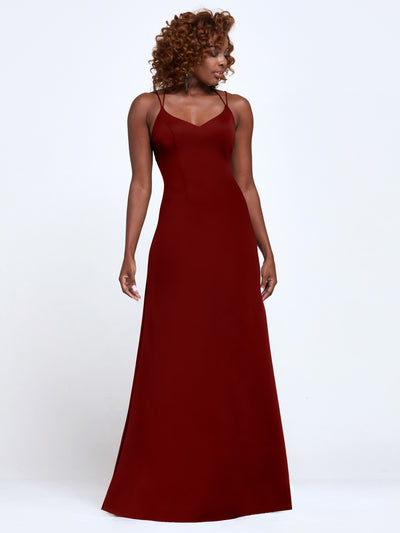 A woman with curly hair wears Avery Austin’s floor-length burgundy A-line dress (style 83045) with spaghetti straps and a fitted bodice, standing against a plain white background.