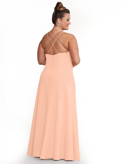 A woman models Avery Austin's 83045: a peach, floor-length A-line dress with delicate cross-back spaghetti straps, her hair in a high bun as she poses against a plain white background.