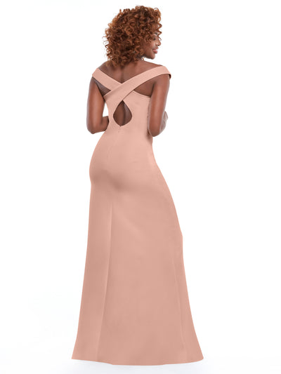 A woman with curly hair models Avery Austin's 83052—a long blush-pink sheath dress with a fitted silhouette, cross-back straps, small keyhole cutout below the shoulders, and a subtle thigh-high side slit—against a white background.