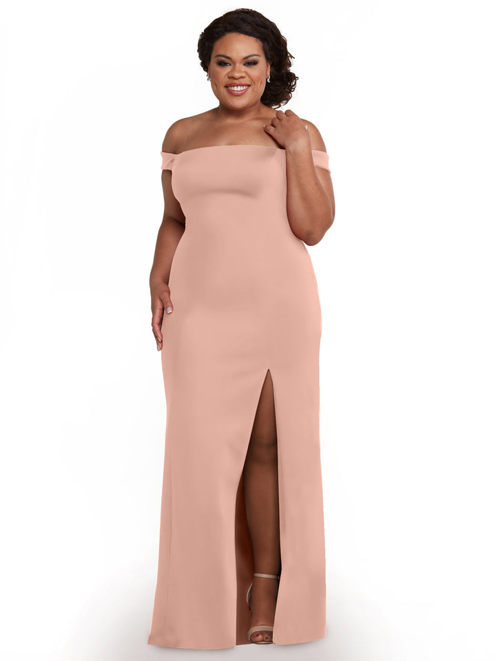 A woman stands smiling against a white background, wearing Avery Austin style 83052, a mauve off-the-shoulder bridesmaid dress with a thigh-high side slit, and touches her hair.