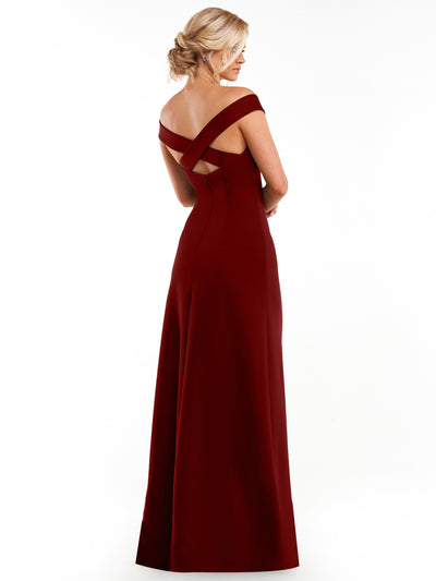 A woman with blonde hair in an updo models Avery Austin’s 83055—a floor-length, off-the-shoulder dark red sheath dress with a crisscross back—against a plain white background.