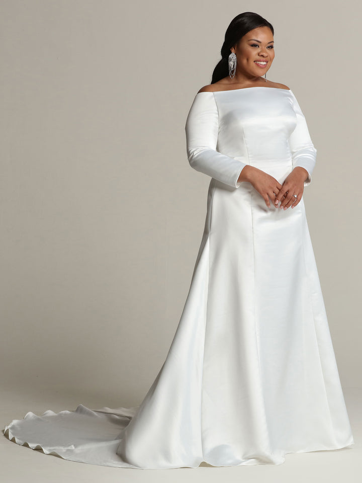 A woman smiles against a plain background, wearing the Ellie wedding dress by Avery Austin—an ivory, long-sleeve, off-the-shoulder satin gown with a small train.