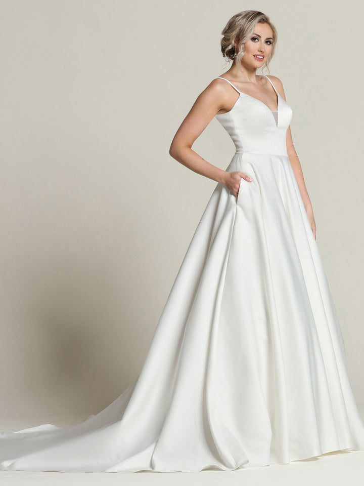 A woman in the Melanie gown by Avery Austin, an ivory satin wedding dress with a fitted bodice, thin straps, V-neckline, and long flowing skirt with pockets, stands against a plain light background showcasing its timeless elegance.
