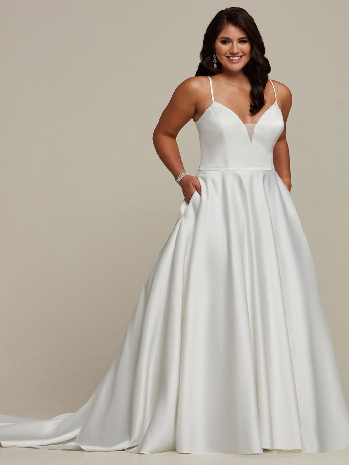 A woman wears the Avery Austin "Melanie" wedding dress in ivory—a sleeveless satin ball gown with thin straps, deep V-neckline, full skirt with pockets. She smiles with one hand in her pocket against a neutral background.