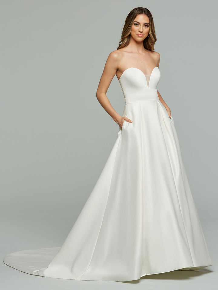 A woman models the Avery Austin "Daisy" wedding dress in ivory, featuring a strapless satin sweetheart neckline, fitted bodice, and full A-line skirt with pockets. She stands against a plain gray background, smiling slightly at the camera.