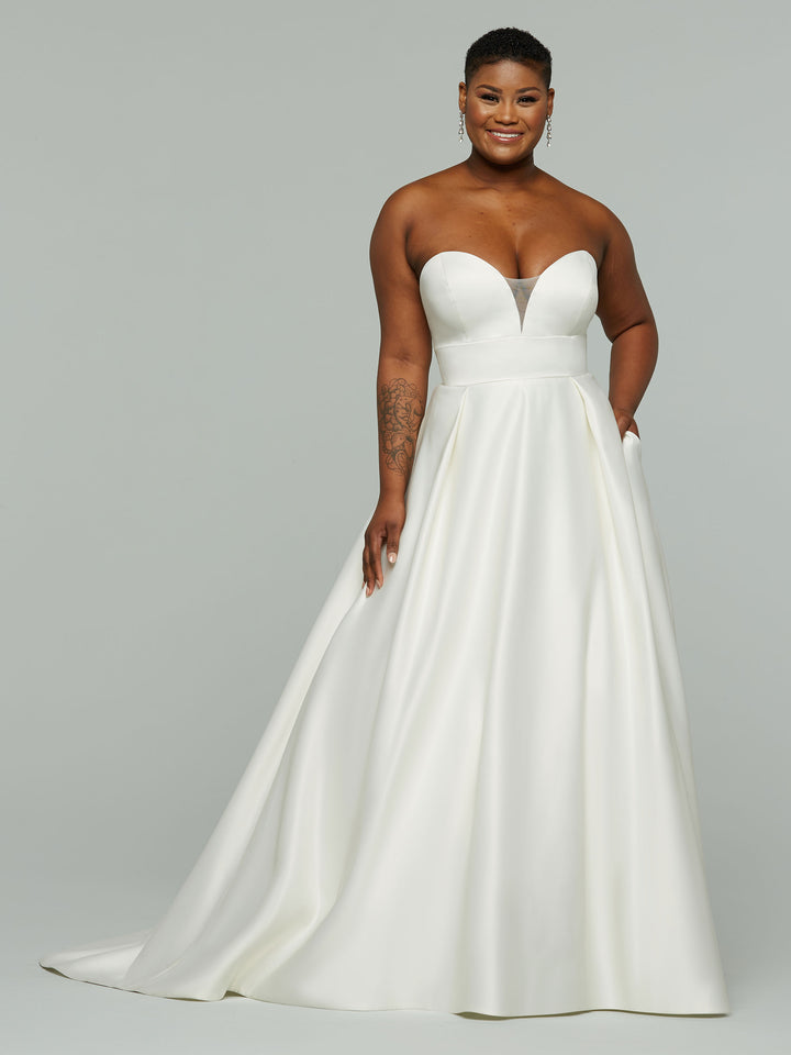 A woman smiles with one hand in her pocket, modeling the Avery Austin "Daisy" strapless ivory satin wedding dress. The gown features a sweetheart neckline, fitted bodice, and A-line silhouette against a plain light gray background.