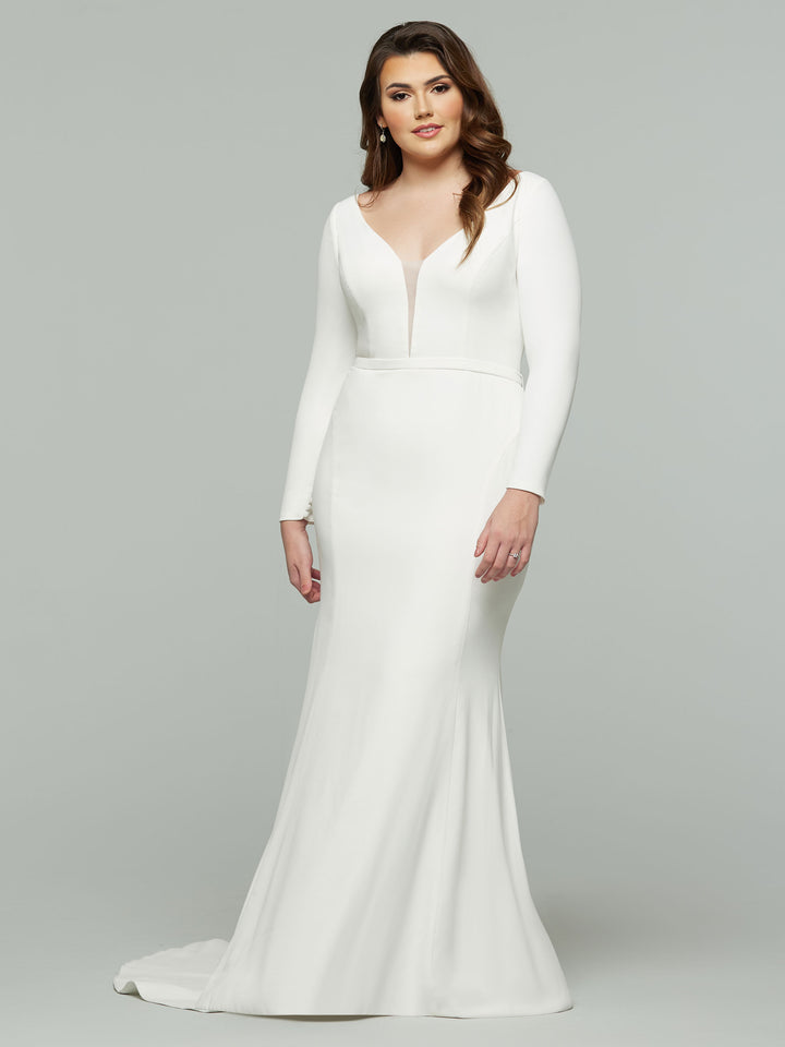 A woman wears the Avery Austin "Felicity" wedding dress in ivory, featuring a fitted silhouette, long sleeves, an illusion plunging v-neckline, and a slight train as she poses against a plain light gray background.