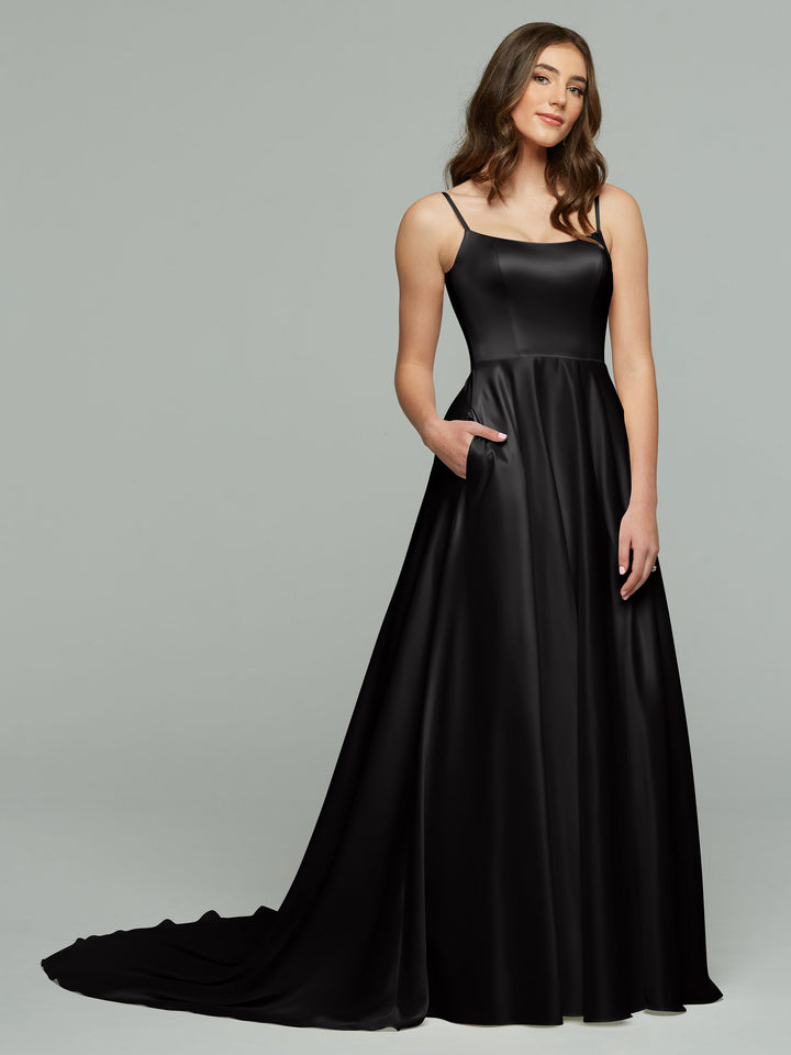 A woman wears the Giselle evening gown by averyaustin in black, featuring thin straps and pockets. She stands against a plain gray background, looking to the side with a soft smile.