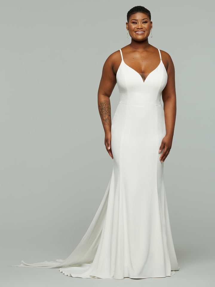 A person smiles in the Avery Austin Harlow—a sleeveless, floor-length ivory wedding dress with thin straps and a V-neckline—standing against a light gray background.