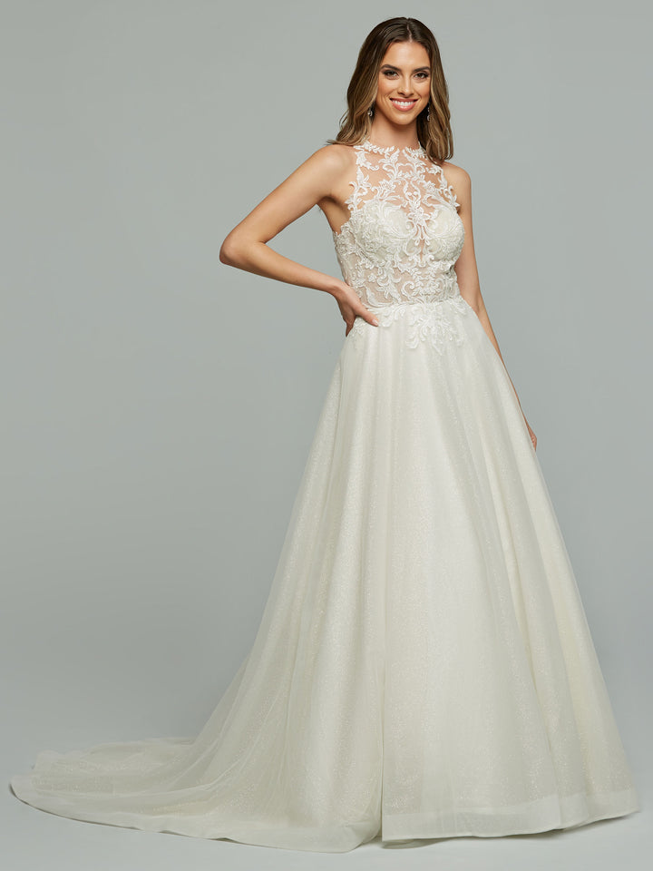 A woman in the Avery Austin "Jamie" ivory wedding dress, featuring a lace bodice, high neckline, fitted waist, and flowing floor-length skirt with a subtle train, smiles as she poses with one hand on her hip.